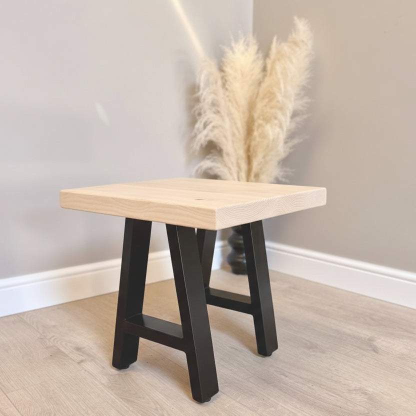 Wooden stool with black legs against a plain wall with pampas grass.