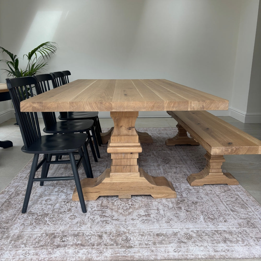 A rustic wooden dining table with a matching bench and black chairs, set on a distressed area rug in a bright room.