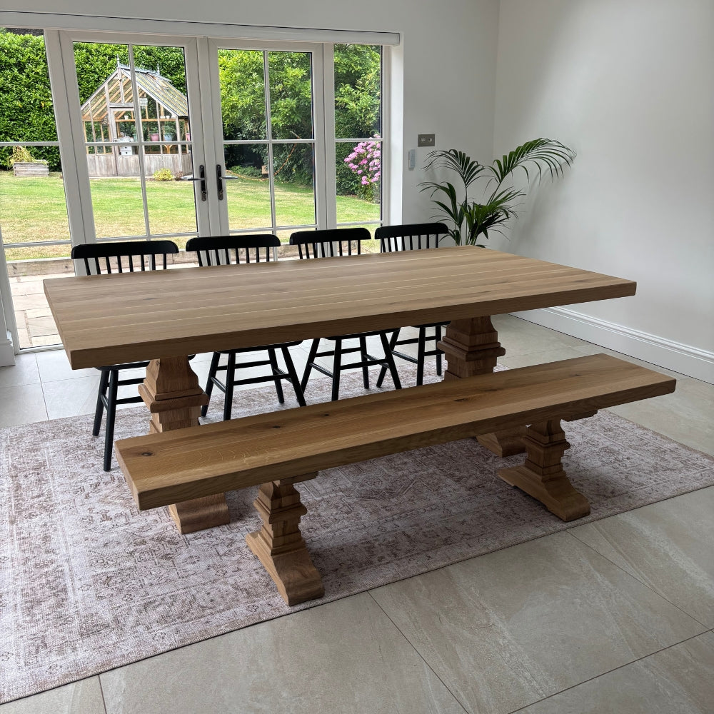 A large rustic wooden dining table with matching bench and black chairs, set in a bright room with white walls, tiled floors, and a window looking out to a garden.