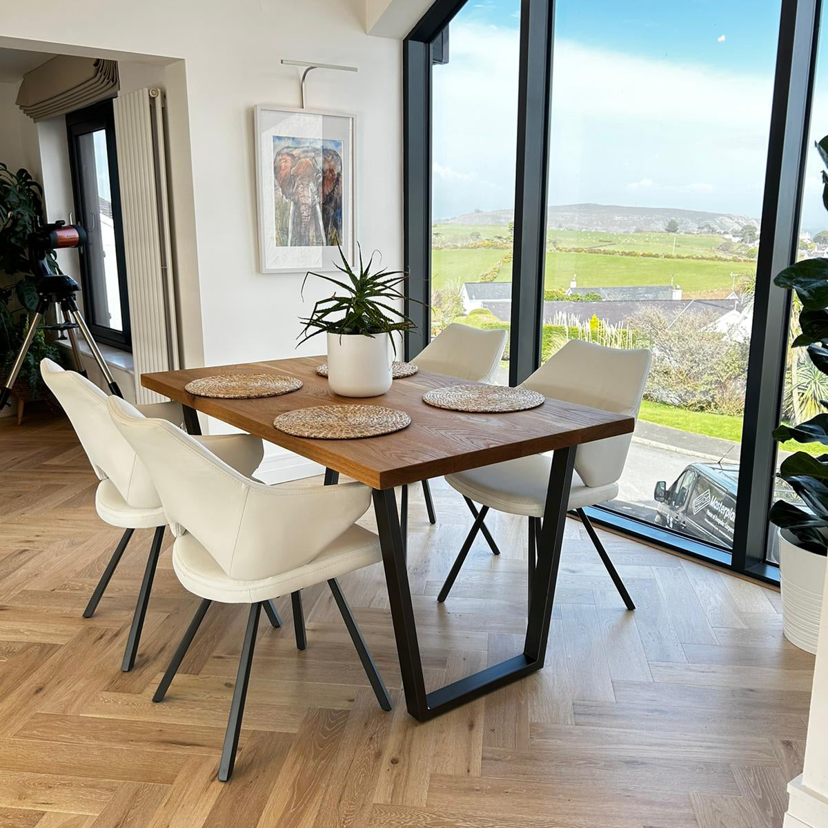 Dining area with wooden table and white chairs in a room with large windows showing a view of greenery.