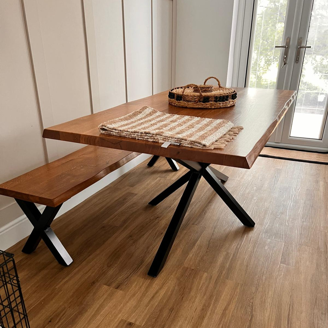 Wooden SOLID OAK WANEY EDGE dining table with bench and woven basket in a room with large windows.