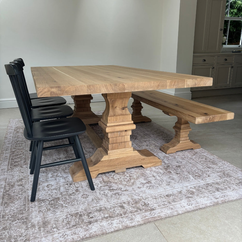 A large rectangular wooden dining table with a trestle base, accompanied by a matching wooden bench and four black spindle-back chairs, set on a distressed beige rug in a bright room with light-colored tiled floors.