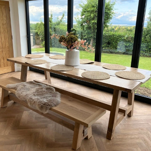 Wooden dining table with benches and placemats in a room with large windows showing a garden view.